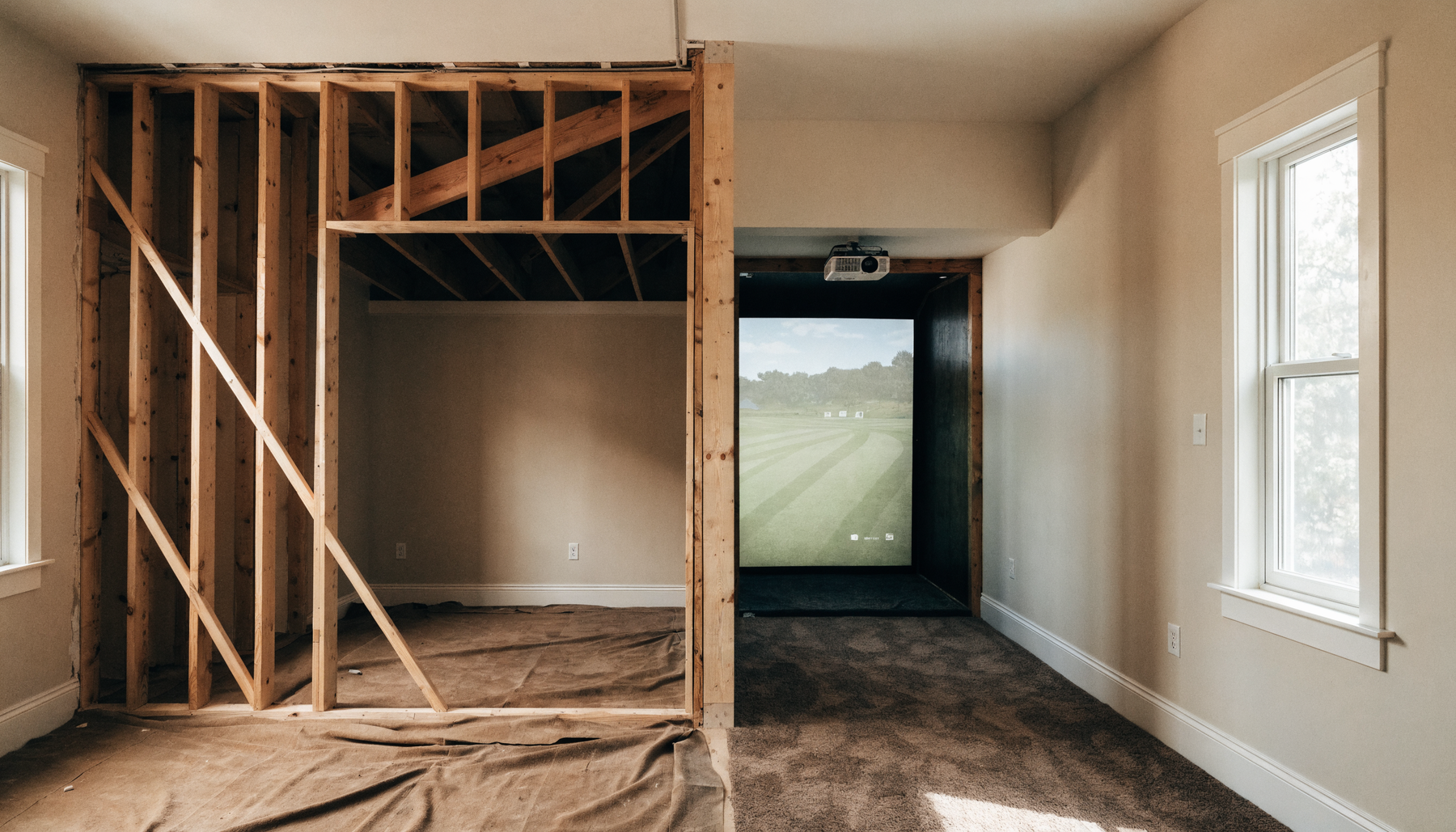 Room mid-remodel, unfinished framing on one side and a finished golf simulator bay on the other
