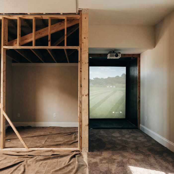 Room mid-remodel, unfinished framing on one side and a finished golf simulator bay on the other
