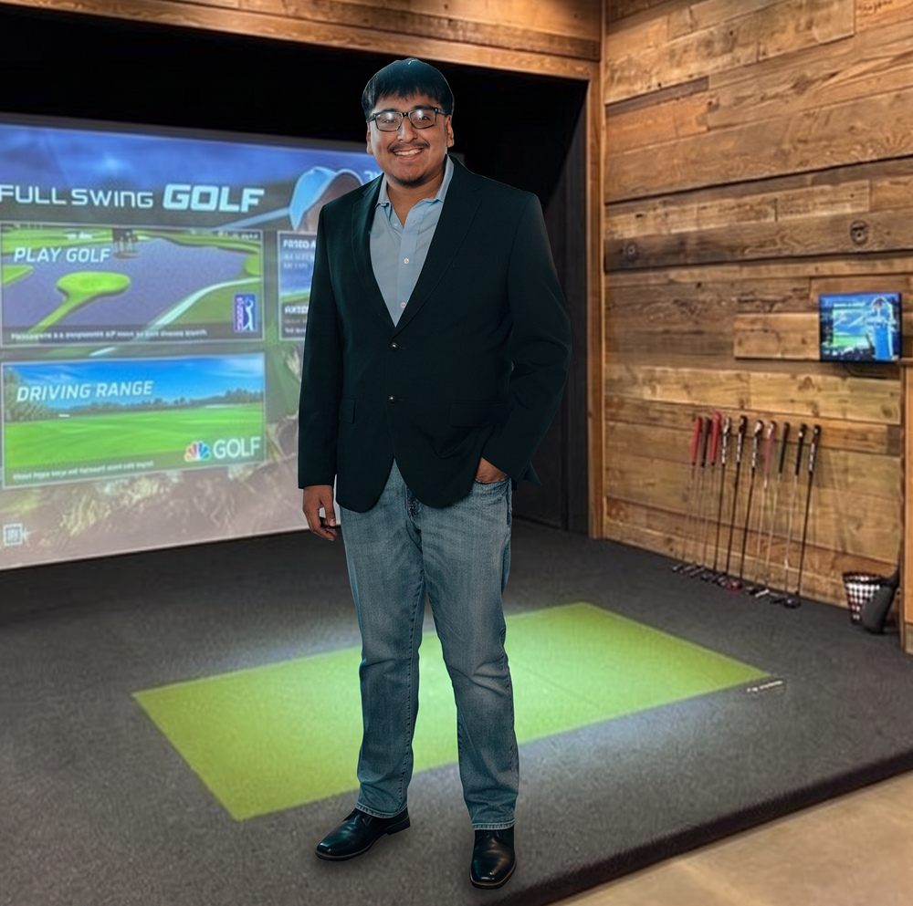 A man wearing a black blazer, light blue shirt, and jeans stands in a home golf simulator studio with wood-paneled walls, a projector screen showing a golf course, and golf clubs hanging on the wall.